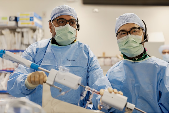 Two clinical team members in procedural attire work together during a hospital procedure while handling medical equipment.